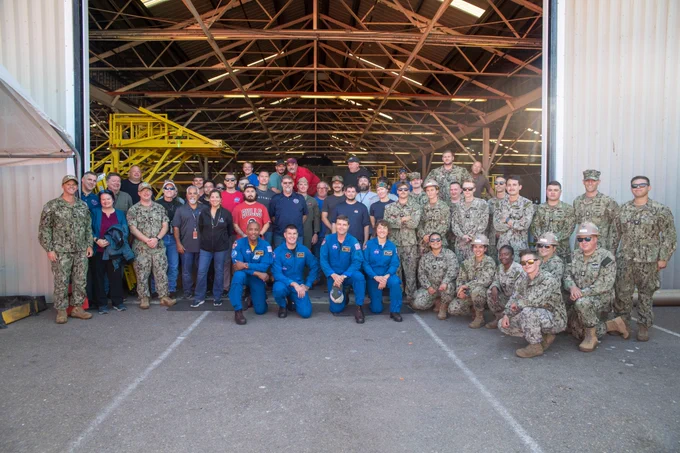 Blue-suited Artemis 2 astronauts kneel in front of a group photo, joined by teams from NASA’s Exploration Ground Systems Program and U.S. Navy sailors from Amphibious Construction Battalion 1.