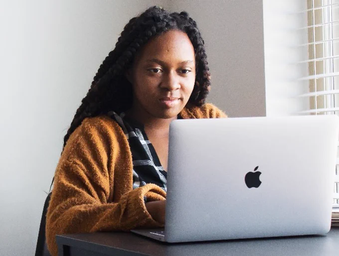 Black woman sitting at a table, looking at her Apple laptop.