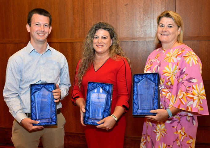 Three adults hold awards and smile. 