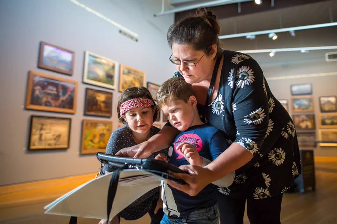 Photo of a white, middle aged woman with tied back brown hair and glasses. Holding a tablet in front of two white children. One is a boy with light brown hair wearing a graphic t-shirt and the other is a girl with a red headband. They are stood in front of a museum exhibition.