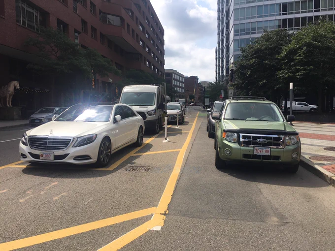 Two columns of cars are parallel-parked, facing the same direction, on a city street. The leftmost column is parked in the car parking lane between the automobile lanes and the bike lanes. The rightmost column is parked within the bike lanes.