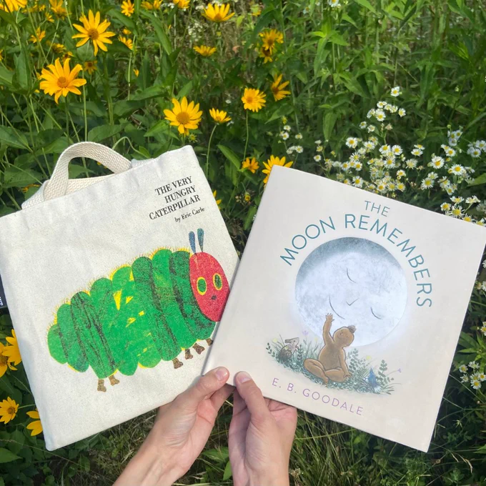 Two hands are holding a Very Hungry Caterpillar tote and the book The Moon Remembers. Flowers are in the background. 