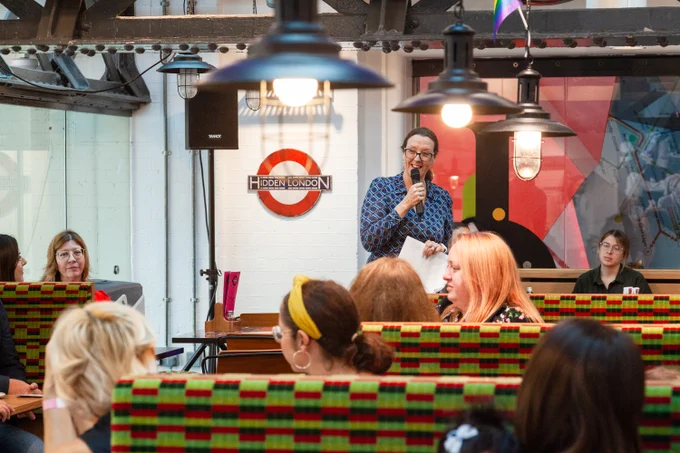 Kids in Museums Family Café Standard Launch, showing Vanessa Eke speaking with a microphone in the centre, a Hidden London logo behind, with people sitting in booths in the foreground and lanterns above.