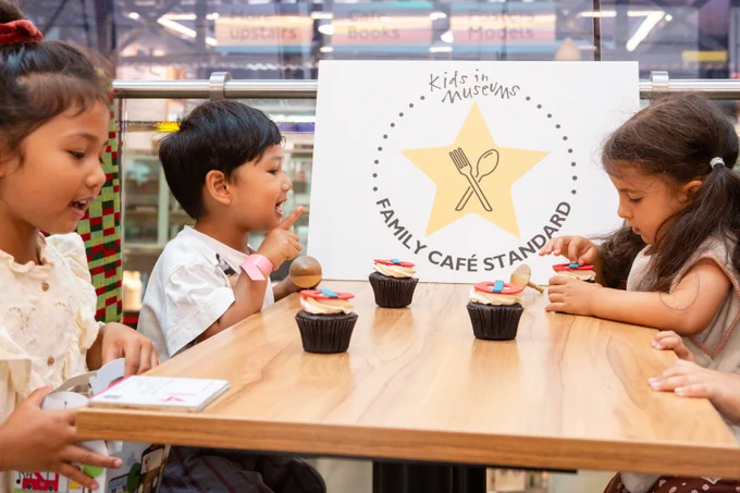 Photograph of the Kids in Museums Family Café Standard Launch, showing three children sitting at a table in a booth, with Tube logo-topped cupcakes on the table and a sign saying 'Kids in Museum, FAMILY CAFÉ STANDARD' behind.