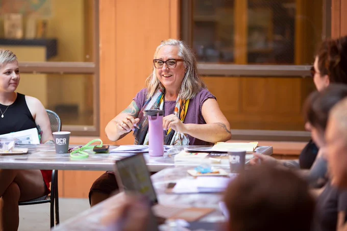 Acclaimed nonfiction writer Barrie Jean Borich shares a laugh with students in an outdoor RWW class she is teaching. Borich is wearing a bright, multi-colored scarf and matching purple shirt and glasses. She is smiling as is a student sitting to her right.