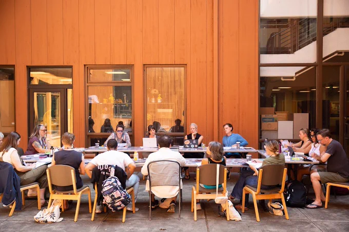 A MFA writing class of 15 people sit with their tables arranged in a rectangle in the outdoor common area in the middle of the AUC. Water bottles, notebooks, books, and laptop computers cover the tables.