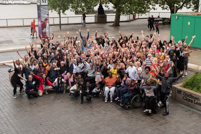 A large group of people stand in a huddle on the pavement by a river. They all put their hands up in celebration. The photo is taken from above.
