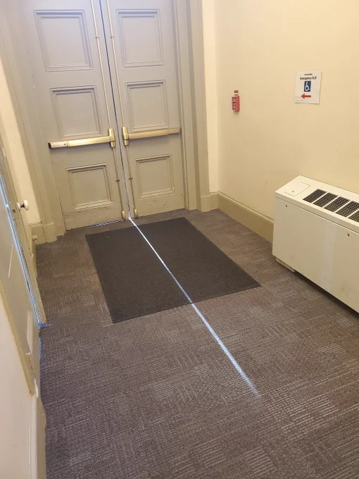A set of green-gray double doors are at the end of a yellow hall with gray carpet. A shaft of light peeks through between the doors on the floor.