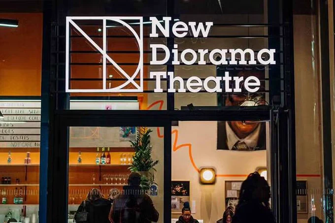 The main entrance to New Diorama Theatre, photographed at night. A person is walking past, inside people sit, while others order drinks at the bar. (Photo credit: Rebecca Need-Menea)