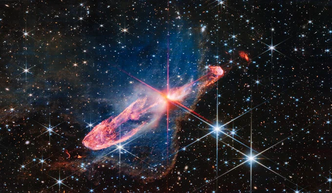 At the centre is a thin horizontal orange cloud tilted from bottom left to top right. It takes up about two-thirds of the length of this angle, but is thin at the opposite angle. At its centre is a set of very large red and pink diffraction spikes in Webb’s familiar eight-pointed pattern. It has a central yellow-white blob, which hides two tightly orbiting stars. The background is filled with stars and galaxies.