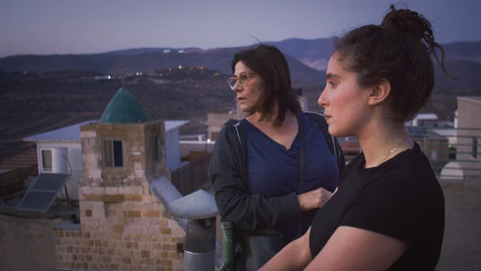 A film still from Bye Bye Tiberias, directed by Lina Soualem. A mother and daughter are standing on a balcony, with serious expressions as they look down below. In the background, buildings and mountains are visible.