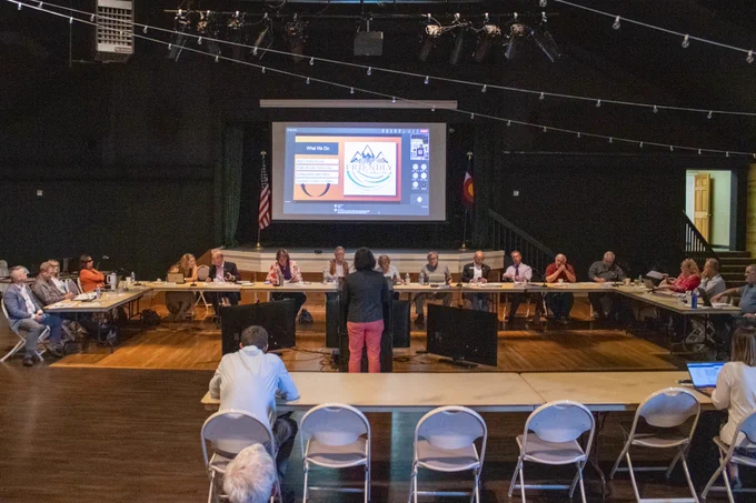 The Pikes Peak Area Council of Governments' Board of Directors sit around a U-shaped table while a person presents at a podium.