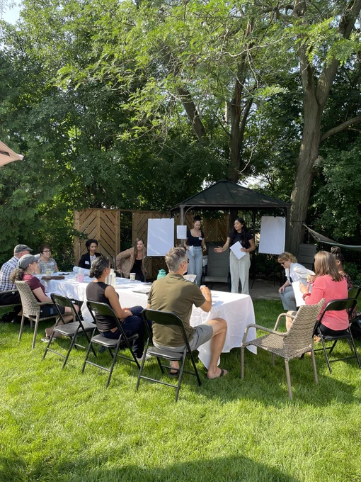People sitting around a table on the grass with two people standing next to two whiteboards, facilitating the conversation.