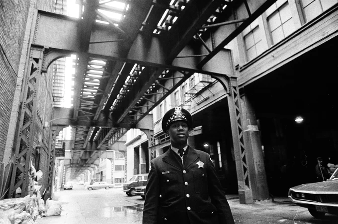 Black & white photograph showing elevated train tracks above an alleyway. In the foreground is a Black male police officer in uniform.