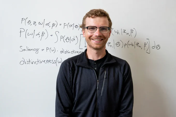 Fletcher Ouren, a red-haired master’s student in the Department of Systems Engineering, stands confidently in front of a whiteboard. He wears horn-rimmed glasses, a dark shirt, and a sweater. With a smile, he poses beside a complex math problem featuring words like "saliency" and "distinctiveness."