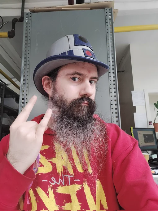 Selfie of myself, a white Non-binary man person with white skin in a red NaztheNeko hoodie and wearing a R2D2 hat. And also having a full grown beard and throw up the devil horns. 