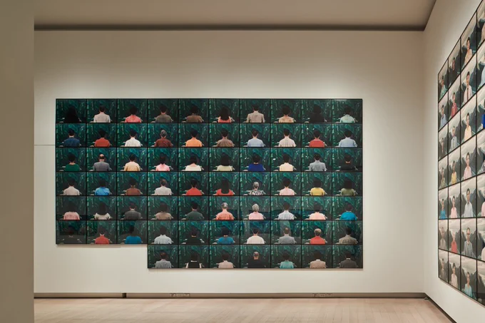 A photograph of a large grid-like art piece with green backgrounds mounted on a gallery wall. 