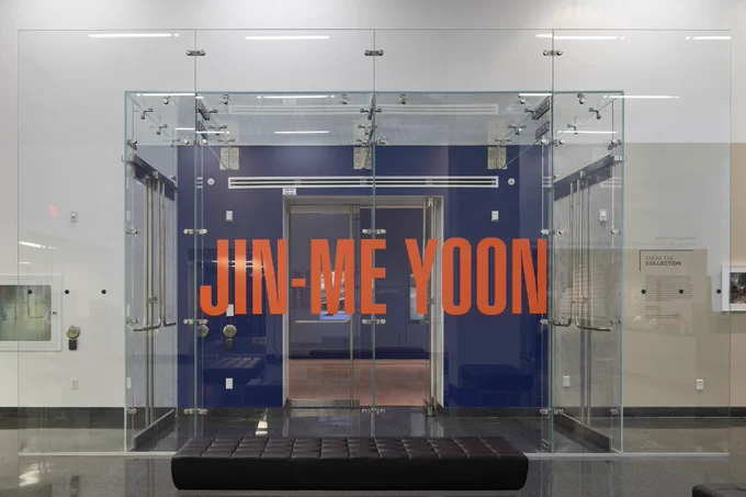 A photograph of the entrance to Jin-me Yoon’s exhibition, the glass entrance reads Jin-me Yoon in bold orange lettering. 