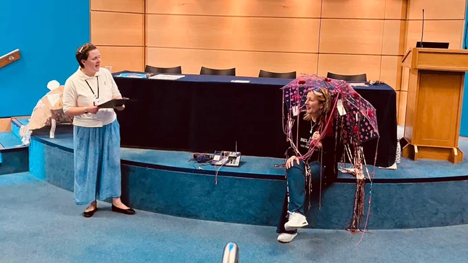 Deborah and Gemma on a stage, presenting at a conference.  Deborah is sitting under a decorated umbrella.
