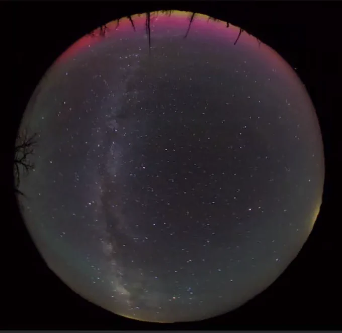 A circular image of the night sky with many stars and the Milky Way visible and bright purple, red, and yellow light of the aurora borealis to the north and a band of green airglow near the horizon.