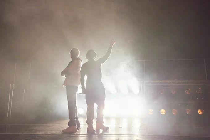 Two hip hop superstars, Tambo and Bones, stand in silhouette, with bright lights from their concert behind them. They form an emblem for all time.