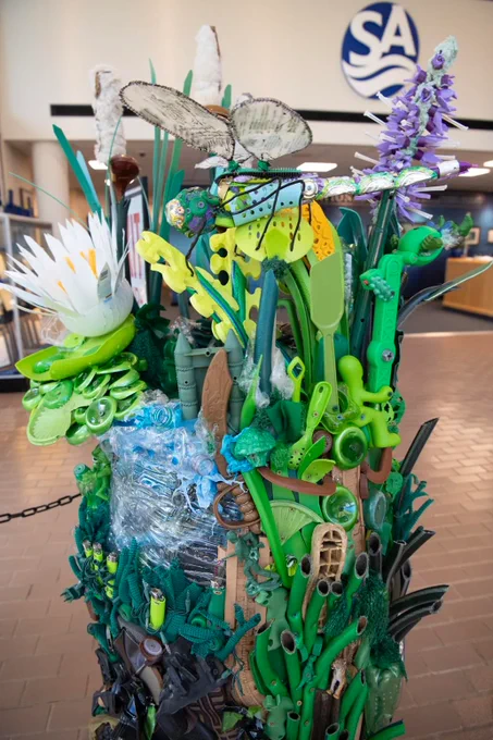 A colorful, large sculpture featuring a dragonfly, grasses, water and a lotus. The sculpture is made of items like garden hoses, plastic bottles, toys and shoes. 
