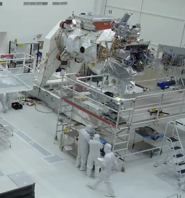 Three engineers in head to toe white coveralls work on the Europa Clipper spacecraft in a very white and sterile looking clean room.