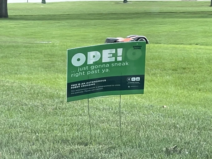 Green lawn sign that reads: “Ope! … just gonna sneak right past you.” In the background is an autonomous robot lawn mower.