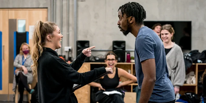 Image shows two people facing each other with theatrical expressions, and other people smiling in the background.