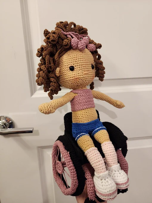 A crochet doll with brown curly hair and medium tan skin. She is seated in a crochet black and pink wheelchair. 