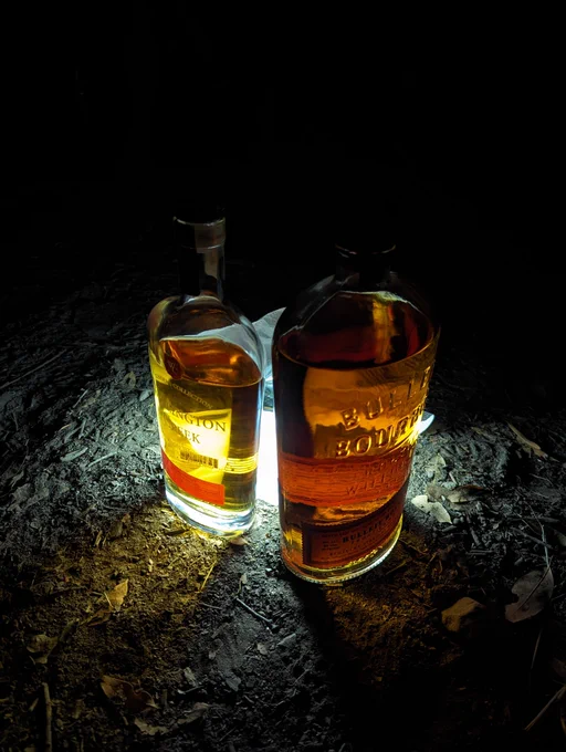 A large bottle of Bulleit bourbon next to a bottle of Wellington Creek bourbon in front of an LED lantern on the ground. It's very dark and the light is shining and diffused through the bottles.