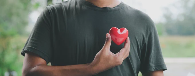 A black man (face not seen) wearing a black T-shirt holds a small shiny red heart to his chest 
