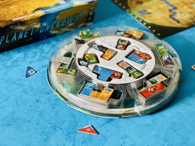 A plastic circular dome is in the centre of the table. Several polyomino holes are cut out, with equivalently shaped cardboard pieces placed in them in piles.  Each piece is split into two colours - red, grey, gold, blue, black and green. In the background is the planet unknown box