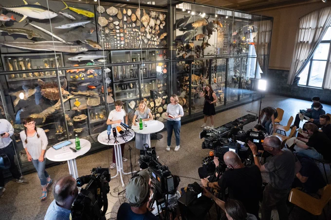 Pressekonferenz in einem Raum des Berliner Naturkundemuseums mit einer Wand mit fossilen Ausstellungsstücken hinter den Sprechenden