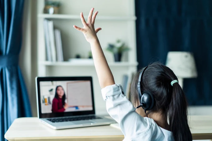 A student raises her hand as a teacher stands in front of a whiteboard on her laptop screen. 