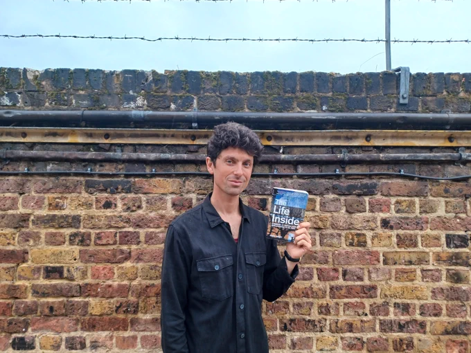 Author and philosopher, Andy West, after delivering a Readconnect workshop at HMP Swaleside. He is holding his book Life Inside and is smiling to camera. He is wearing a black shirt and is standing in front of  brick wall.