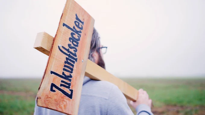 Eine Person steht auf einem Feld und trägt ein Holzschild über der Schulter mit der Aufschrift: Zukunftsacker.
