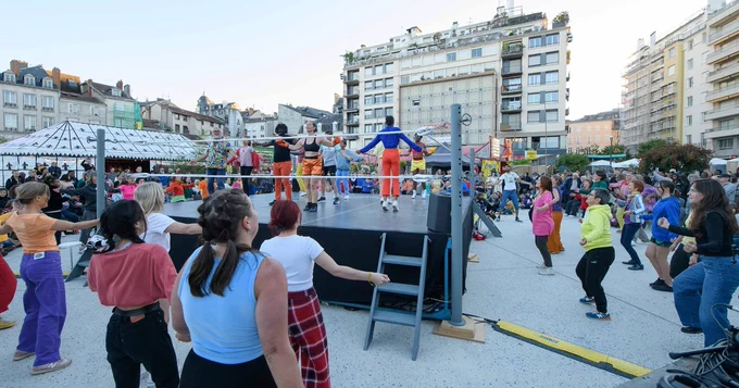 A group of people are outside, surrounding and on a wrestling stage. They seem to be dancing, following the instructions of someone standing on the stage. 