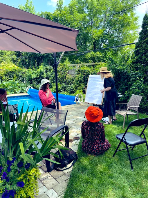 Members of the Co-Design Hub engage in prototype building using a large easel with white paper and markers to brainstorm ideas as a group. The session is outside, surrounded by greenery and a pool.