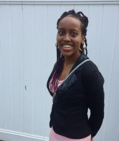 A dark-skinned woman stands in front of a white fence