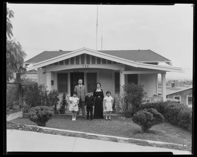 Black and white photograph of a family standing in front of a house. There is a father, mother, and three young children.