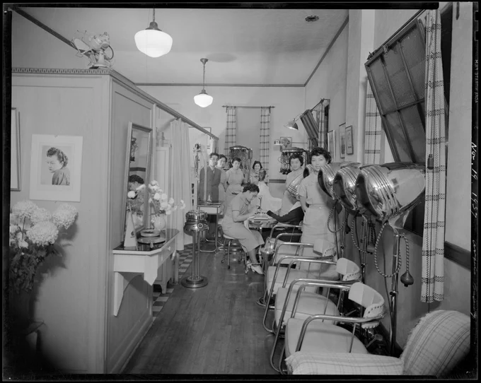 Black and white photograph of the inside of a hair salon. Several women are standing at the back of the salon looking at the camera. There are chairs and hair dryers in the salon.