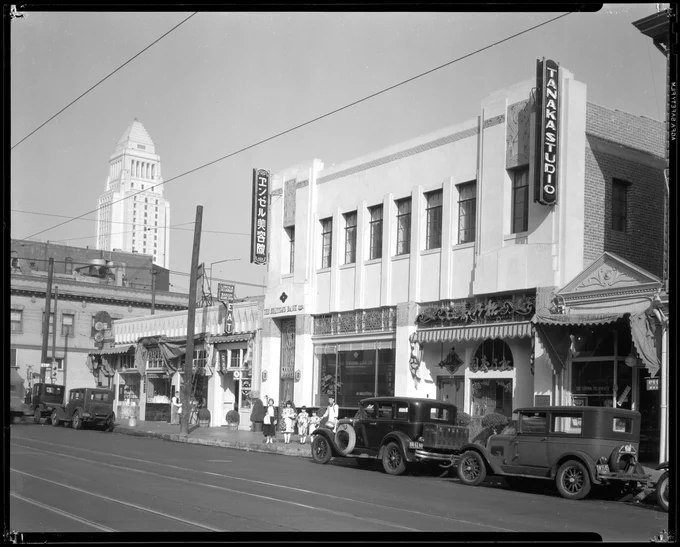Black and white photograph of a street view of Little Tokyo, Los Angeles. City Hall is seen in the background. Cars are parked along the side of the street and a sign on the building reads, "Tanaka Studio"