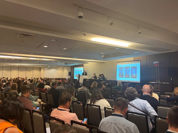Conference room with 100  people listening to a panel discussion on an elevated stage, with two screens at the front of the room. 