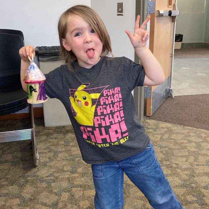 Child with brown hair, jeans and a gray Pokemon t-shirt holds up a hand-painted birdhouse, while sticking out their tongue.