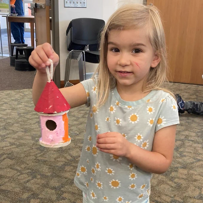Child with blonde hair and a gray t-shirt with daisies on it holds up a hand-painted birdhouse.