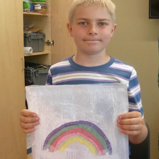 Child with striped t-shirt smiles and holds up a hand-made suncatcher with a rainbow on it