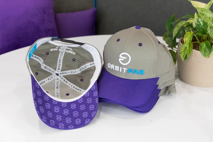Picture of Orbit Fab hat outside and inside. The hat has a purple bill and grey body, with the Orbit Fab logo embroidered on the front. Inside the hat, hem tape reads build - break - learn - fly.