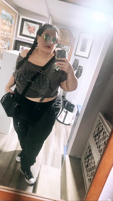 Marianne, a very fat pale femme, leans forward into a mirror to take a selfie. She’s wearing black sweatpants, a grey tshirt tied up to show her midriff, and black converse with moons and stars on them. She’s got on red lipstick, a black crossbody jelly basket bag, and mirrored aviators shaped like hearts and her hair is in braided pigtails.