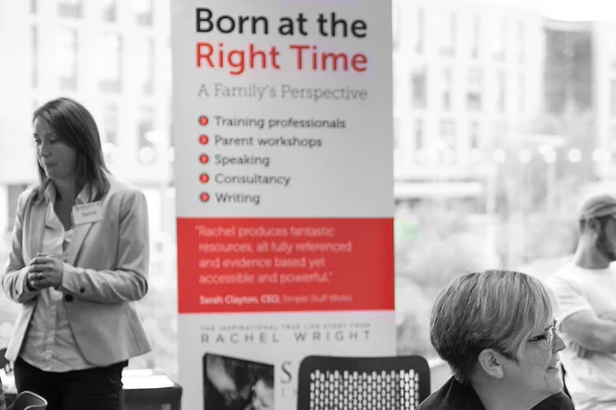 Black and white photograph of a professional lady with shoulder-length hair wearing a blazer and shirt next to a training banner from Born at the Right Time. The banner has highlights picked out in red.